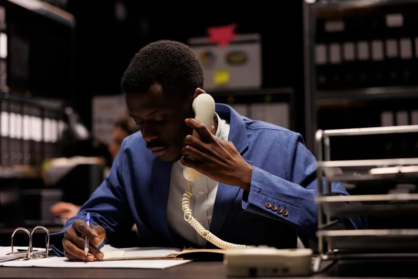 Менеджер-афроамериканец разговаривает по стационарному телефону в складском помещении, обсуждая с удаленным бухгалтером исследование по менеджменту. Бизнесмен поздно ночью работает над бухгалтерским отчетом.